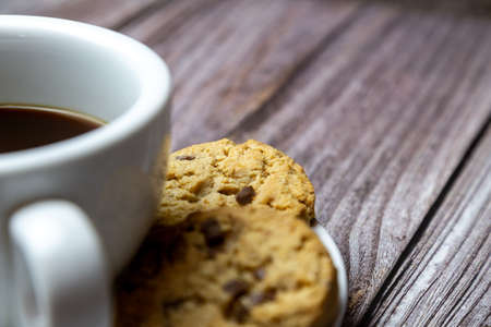 chocolate chip cookies next to a cup of coffee on a saucer on a rustic wooden tableの写真素材