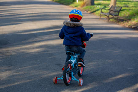 A small boy wearing a cycle helmet riding a bike in the park with stabilisers or stabilizersの写真素材