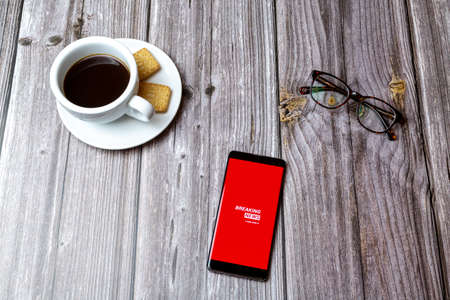 03-03-2021 Portsmouth, Hampshire, UK A mobile phone or cell phone laid on a wooden table with the Breaking news app open next to a coffeeのeditorial素材