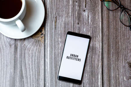 02/10/2021 Portsmouth, Hampshire, UK A mobile phone or cell phone laid on a wooden table with the Urban Outfitters app open on screen next to a coffeeのeditorial素材