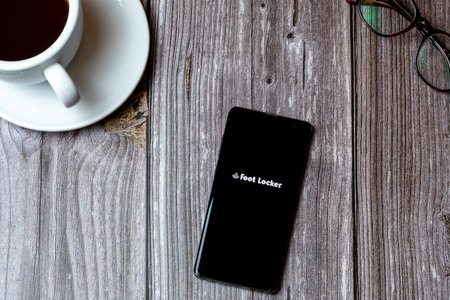 02/10/2021 Portsmouth, Hampshire, UK A mobile phone or cell phone laid on a wooden table with the Foot locker app open on screen next to a coffeeのeditorial素材