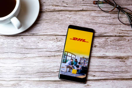 02-08-2021 Portsmouth, Hampshire, UK A mobile phone or cell phone laid on a wooden table with the DHL delivery app open next to a coffee and glassesのeditorial素材