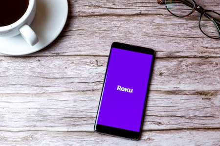 02-08-2021 Portsmouth, Hampshire, UK A mobile phone or cell phone laid on a wooden table with the Roku TV app open next to a coffee and glassesのeditorial素材