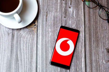 02/10/2021 Portsmouth, Hampshire, UK A mobile phone or cell phone laid on a wooden table with the Vodafone app open on screen next to a coffeeのeditorial素材