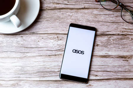 02-08-2021 Portsmouth, Hampshire, UK A mobile phone or cell phone laid on a wooden table with the Asos shopping app open next to a coffee and glassesのeditorial素材