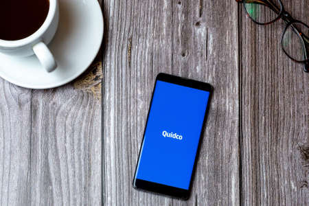 02/10/2021 Portsmouth, Hampshire, UK A mobile phone or cell phone laid on a wooden table with the quidco cashback app open on screen next to a coffeeのeditorial素材