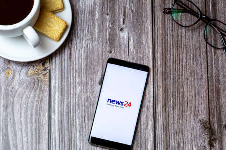 02-24-2021 Portsmouth, Hampshire, UK A mobile phone or cell phone on a wooden table with the News 24 app open next to a coffee and glassesのeditorial素材