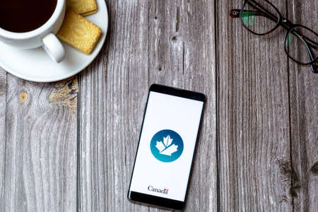 02-24-2021 Portsmouth, Hampshire, UK A mobile phone or cell phone on a wooden table with the Canada Business app open next to a coffee and glassesのeditorial素材