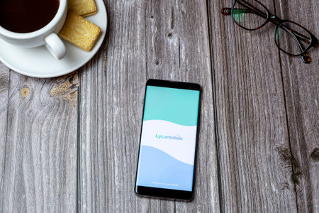 02-24-2021 Portsmouth, Hampshire, UK A mobile phone or cell phone on a wooden table with the Lycamobile app open next to a coffee and glassesのeditorial素材
