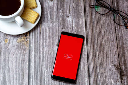 02-24-2021 Portsmouth, Hampshire, UK A mobile phone or cell phone on a wooden table with the Economist app open next to a coffee and glassesのeditorial素材