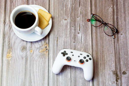 03-23-2021 Portsmouth, Hampshire, UK A Google Stadia controller resting on a wooden table next to a coffee cup and glassesのeditorial素材