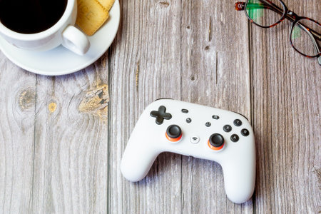 03-23-2021 Portsmouth, Hampshire, UK A Google Stadia controller resting on a wooden table next to a coffee cup and glassesのeditorial素材