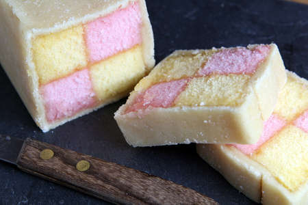Freshly cut Battenberg Cake on dark slate background with knife. Pink and yellow sponge covered in marzipanの写真素材