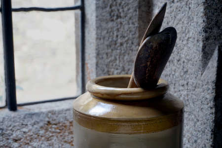 Old wooden cooking spoons in a large ceramic jug or pot, with stone walls and window in backgroundの写真素材