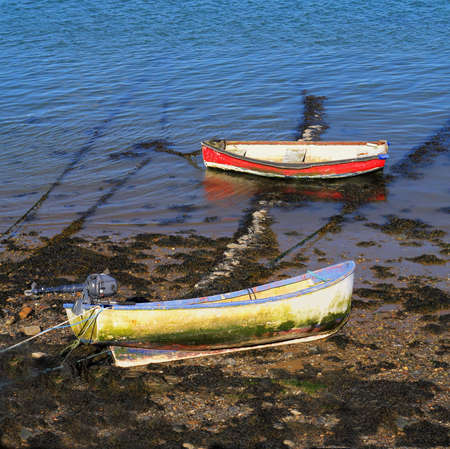 Two old rowing boats, red and yellow, at low tide in a river estuary, blue waterの写真素材