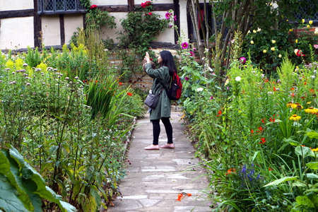 Stratford-upon-Avon, UK - July 21 2017: Chinese female tourist takes a cellphone photograph among pretty flowers in an English country gardenのeditorial素材