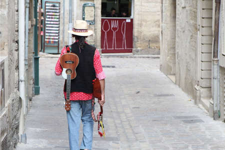 Pezenas, Herault, France - Aug 26 2017: Musician wearing a straw fedora hat, carrying a mandolin and ukelele, walking down a cobbled street towards a small cafeのeditorial素材