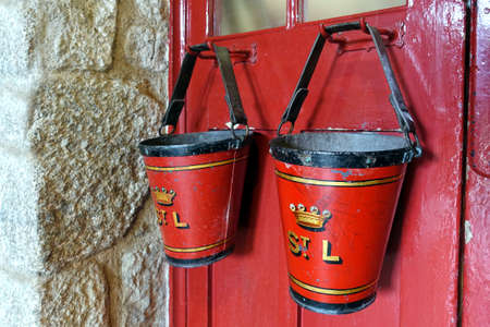 Marazion, Cornwall, UK - April 13 2018: Two old fashioned vintage antique fire buckets hanging on a red doorのeditorial素材