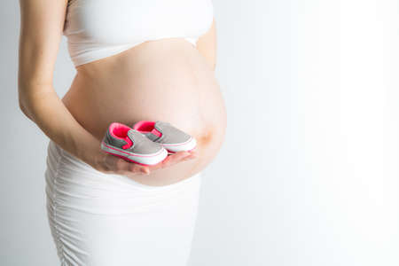 A pregnant woman holds a pair of baby shoes in front of her pregnant belly.の写真素材