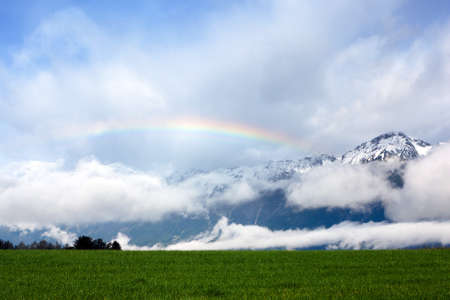 Rainbow over a green meadow in the Alps, with snowcapped hills in the background の写真素材