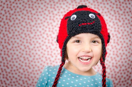 Portrait of little girl smiling and being happy. Child is wearing a woolen cap in the shape of ladybird on a background with red dots. Happy Childrenの写真素材