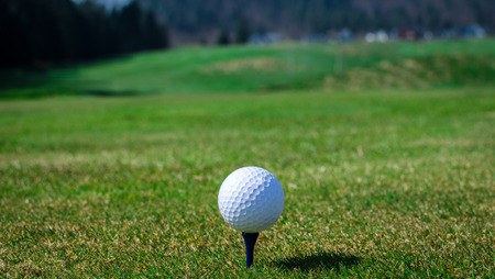 Golf ball on teeing area with green grass ahead and mountains in background. Soft focus or shallow depth of field.の写真素材
