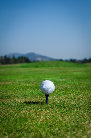 Golf ball on teeing area with green grass ahead and mountains in background. Soft focus or shallow depth of field.の写真素材