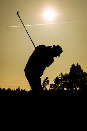 Silhouette of old man playing golf. Senior citizen is wearing a hat and swinging the club with sun and airplane contrails in the background. Also a silhouette of forest and trees in the evening or morning dusk or dawn.の写真素材