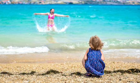 Mother and child having fun at the beach. Little girl is watching mother having fun in the sea. She is jumping and splashing water.の写真素材