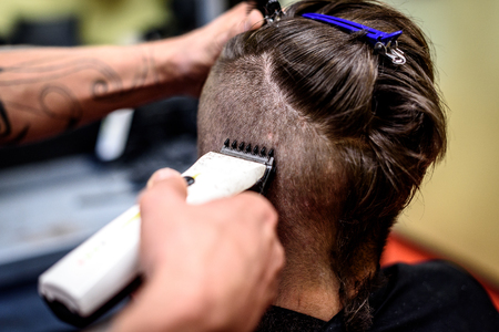 Close-up of a man getting his hair shaved with trimmer. Hairdressers hand is holding electric trimmer and shaving a costumers head.の写真素材