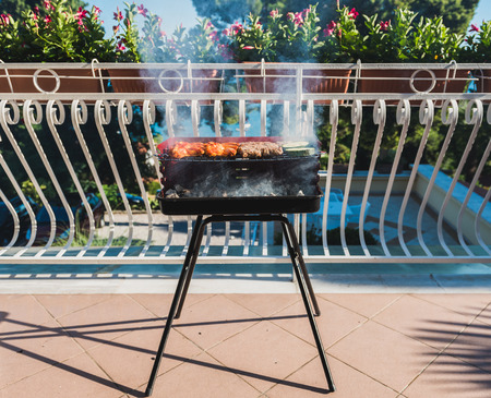 Delicious meat on barbecue grill with coal on balcony. Grilling food on a balcony or a terrace with wonderful view. Small cheap BBQ grill at home.の写真素材