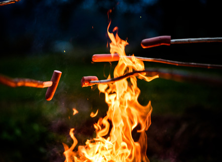 Making and cooking Hot dog sausages over open camp fire. Grilling food over flames of bonfire on wooden branch - stick spears in nature at night. Scouts way of preparing food.の写真素材
