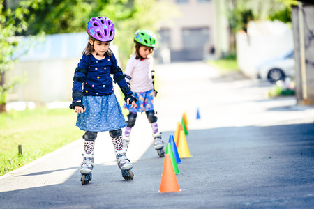 Children learning to roller skate on the road with cones. Twin girls are practising safe roller skating on a home driveway road wearing protective gear - helmets, knee, elbow and hand protectors or pads.の写真素材