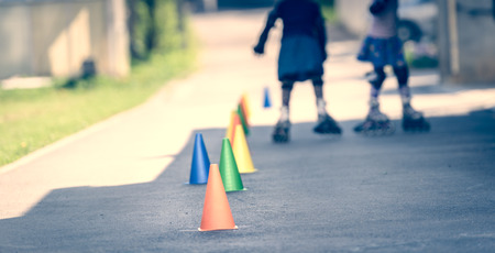 Children learning to roller skate on the road with cones. Twin girls are practising safe roller skating on a home driveway road wearing protective gear - helmets, knee, elbow and hand protectors or pads.の写真素材