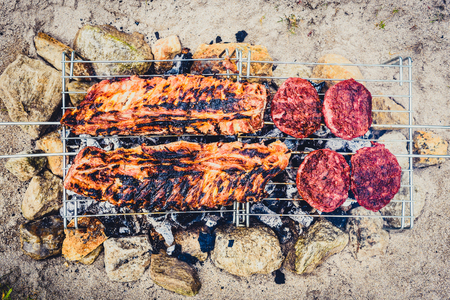 Pork ribs and burgers on homemade improvised BBQ barbecue grill. Making Churrasco  on coal, fire, briquettes BBQ grill in the sand with rocks stones Camping and charcoal grilling ideas life hacks.の写真素材