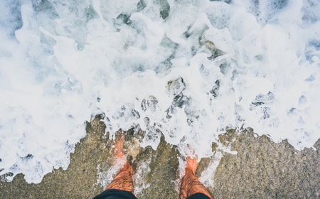 Person standing in ocean sea waves on sandy beach. Barefoot legs soaking in sea water. Man feet standing on sandy beach of Atlantic ocean in Galicia - Spain.の写真素材