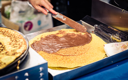 Making of crepes pancakes in open market festival fair. A hand is making crepes outdoors on a metal griddle with wooden stick on a outdoor summer fest.の写真素材