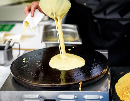 Making of crepes pancakes in open market festival fair. A hand is making crepes outdoors on a metal griddle with wooden stick on a outdoor summer fest.の写真素材