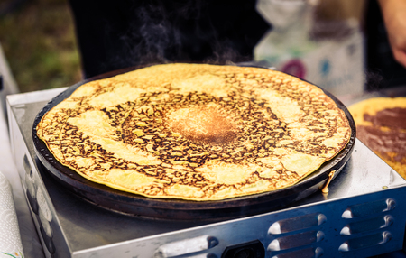 Making of crepes pancakes in open market festival fair. A hand is making crepes outdoors on a metal griddle with wooden stick on a outdoor summer fest.の写真素材
