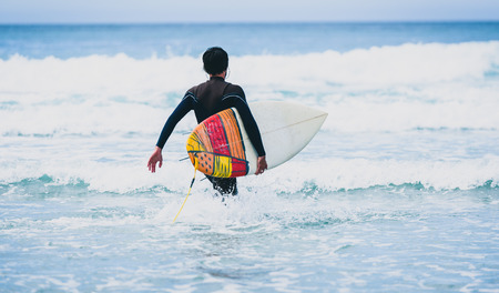 Surfer guy with surfboard in hand running towards big waves. Man in surfing wet suit is running in the waves of cold atlantic ocean in Galicia, Spain.の写真素材