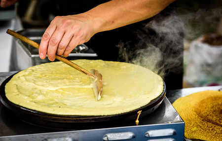 Making of crepes pancakes in open market festival fair. A hand is making crepes outdoors on a metal griddle with wooden stick on a outdoor summer fest.の写真素材