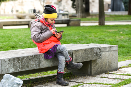 Little girl child using a mobile phone in public park. Instead of playing outside, child is playing games on telephone, sitting in granite bench. Children using social network apps on phone or tablet.の写真素材