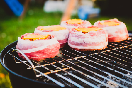 Making home made Beer Can Bacon Burgers on barbecue grill. Preparing stuffed patties, wrapped  in bacon and grilling on indirect heat in nature at back yard BBQ picnic. Low and Slow technique with sunshine in background.の写真素材