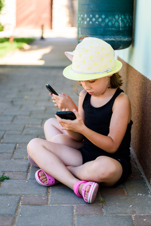 Girl child using smartphone for games or internet on vacations. Little girl is playing with smartphone, sitting on paved floor in the shade, wearing sandals and a hat.の写真素材