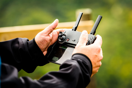 Hand holding drone quadcopter UAV remote control console controls. 
Professional  drone operator is flying a remote control aerial vehicle drone in the nature with green background.の写真素材