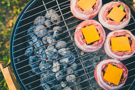 Making home made Beer Can Bacon Burgers on barbecue grill. Preparing stuffed patties, wrapped  in bacon and grilling on indirect heat in nature at back yard BBQ picnic. Low and Slow technique with sunshine in background.の写真素材