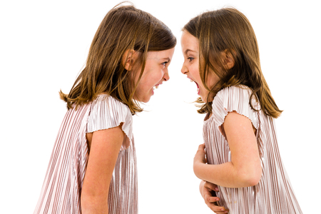 Identical twin girls sisters are arguing yelling at each other. Angry girls are shouting, yelling and arguing with emotional expression on faces. Frontal profile view of children. Isolated on white.の写真素材