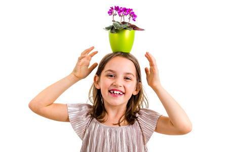 Girl carrying flower pot on head, having fun smiling. Little girl in dress holding green flower pot with viola flowers on her head having fun, goofing around. Studio shot isolated on white backgroundの写真素材