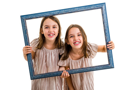 Identical twin girls are making happy expressions with picture frame. Children, sisters, girls posing in studio with picture frame, making different facial expressions. Family portrait, frontal view.の写真素材