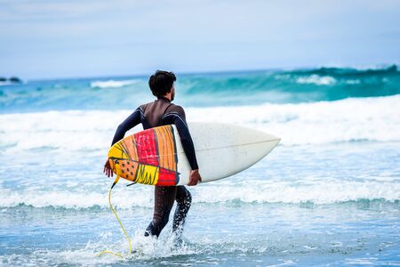 Surfer guy with surfboard in hand running towards big waves. Man in surfing wet suit is running in...